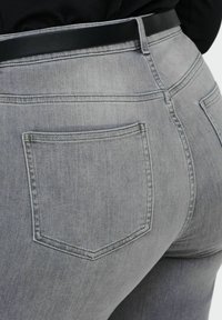 Close-up van een persoon die lichtgrijze spijkerbroek draagt met achterzakken en een zwarte riem zichtbaar bij de taille.