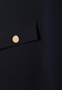 Navy fabric with a smooth texture, featuring a button and flap detail; the button is gold circular metal, positioned on a pocket.