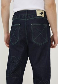 Dark blue denim jeans with green stitching, featuring rear pockets with cross patterns and a tag on the waistband. Exposed seams accentuate design.