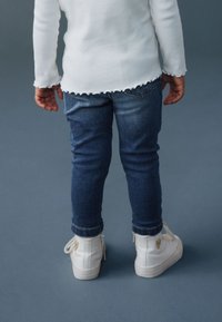 Fehér hosszú ujjú bordázott ing fodros szegéllyel, kék farmernadrág, és fehér magas szárú tornacipő szív részlettel az oldalon.