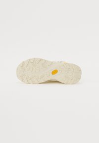 Sko såle i beige med teksturert gummi, med en gul Vibram-logo. Tredesignen viser varierte grep mønstre og små flekker.