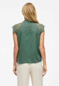 Zielona bluzka z koronkowymi akcentami na ramionach i wysokim dekoltem, z otwarciem z tyłu oraz gładką fakturą materiału.