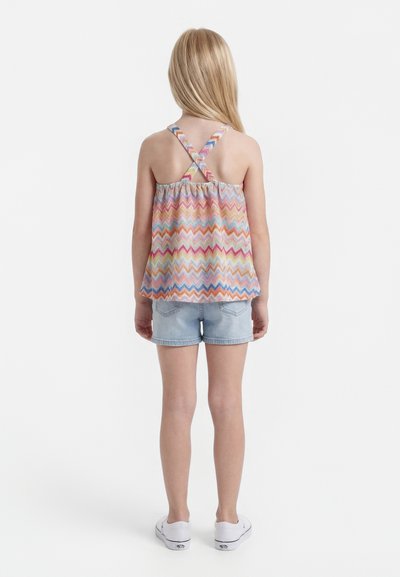 Παιδί με μακριά ξανθά μαλλιά φοράει αμάνικο πολύχρωμο τοπ με ζιγκ ζαγκ σχέδιο και σταυρωτούς ιμάντες, τζιν σορτς και λευκά αθλητικά παπούτσια, στέκεται με την πλάτη προς τα εμπρός.