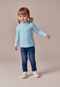 Světlomodrý rolák s dlouhými rukávy z měkké látky, doplněný tmavě modrými džínami a bílými teniskami, na neutrálním pozadí.