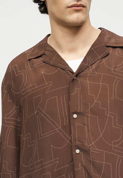 Άνδρας που φοράει καφέ πουκάμισο με κουμπιά, με λευκά αφηρημένα διάστικτα γραμμικά σχέδια και φαρδιά γιακά πάνω από λευκό υποκάμισο.