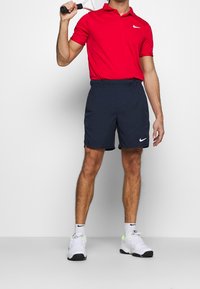 Camiseta polo roja con el logo de Nike, combinada con pantalones cortos azul marino con cinturilla elástica y zapatillas blancas con acentos negros y neón.