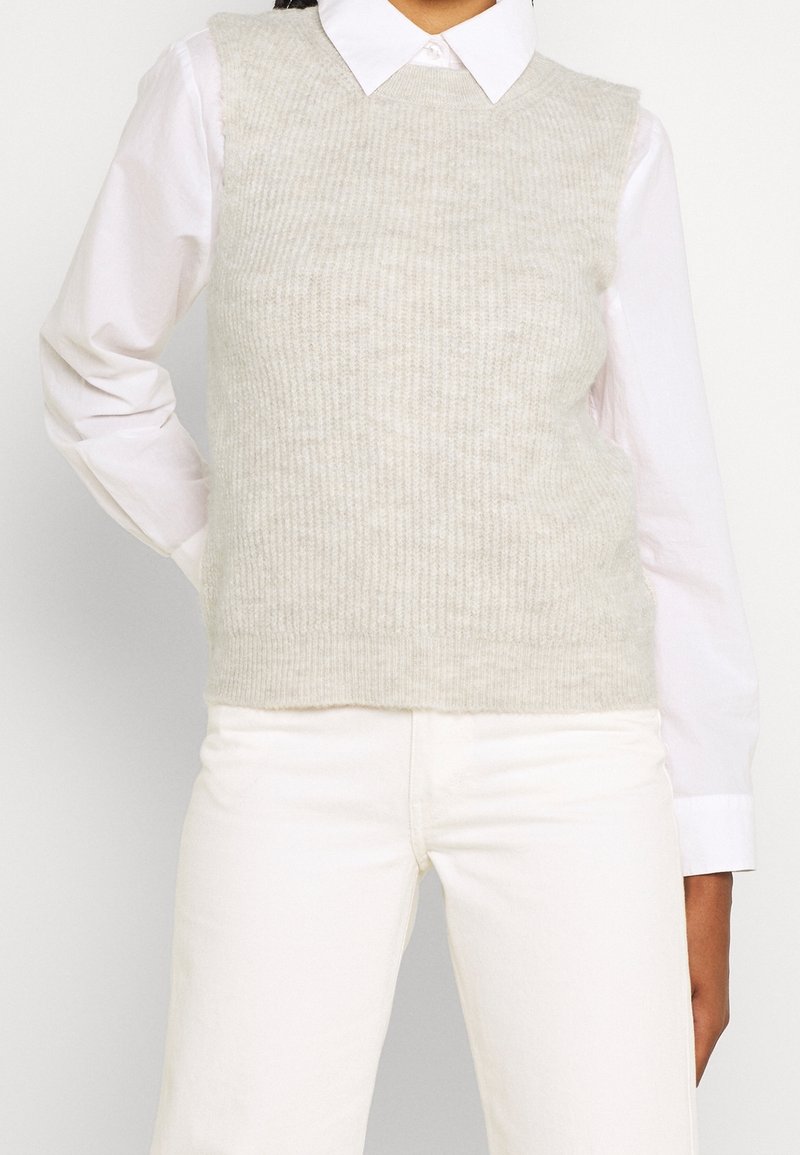 Femme portant un pull sans manches en maille gris clair par-dessus une chemise blanche à col et un pantalon blanc, debout avec une main derrière le dos.