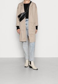 Person wearing beige long coat, black top, light blue jeans, white ankle boots, and black crossbody bag with silver chain detail.