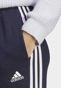 Marinblå joggerbyxor med vit Adidas-logotyp, med tre vita sidostripes och en sidoficka. Texturerade gråa ärmslut på en tröja syns.