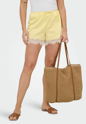 Osoba nosząca bladożółte szorty z koronkową lamówką i brązowe klapki, trzymająca przy boku dużą plecioną słomianą torbę.