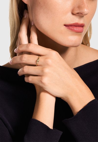 Frau mit hellem Teint und blondem Haar, die ein schwarzes Oberteil trägt und einen goldenen Ring mit Knotendetail an den Fingern ihrer linken Hand zeigt.