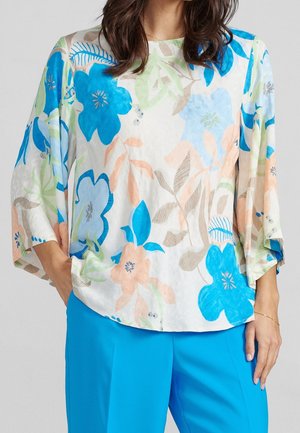 Kvinde iført løs hvid bluse med store blå, fersken- og grønne blomsterprint og lyseblå bukser, hænder i lommerne.