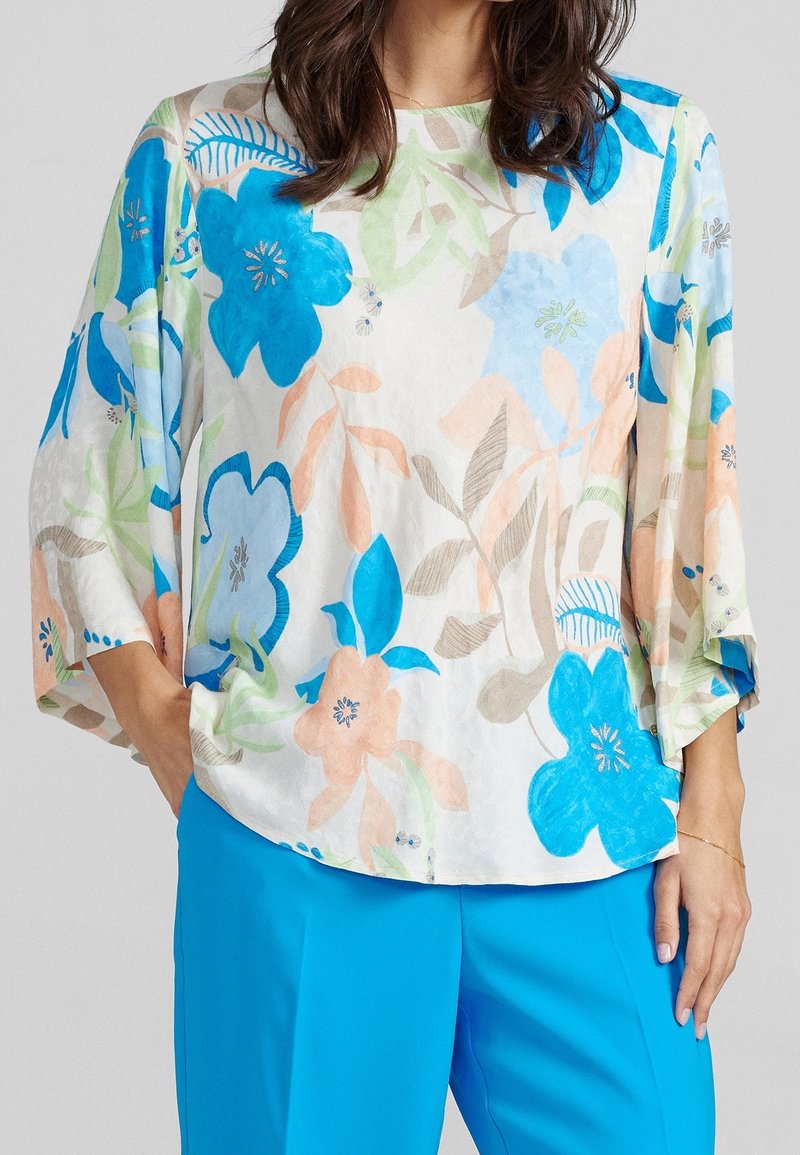 Femme portant une blouse blanche ample avec de grands motifs floraux bleus, pêche et verts et un pantalon bleu vif, mains dans les poches.