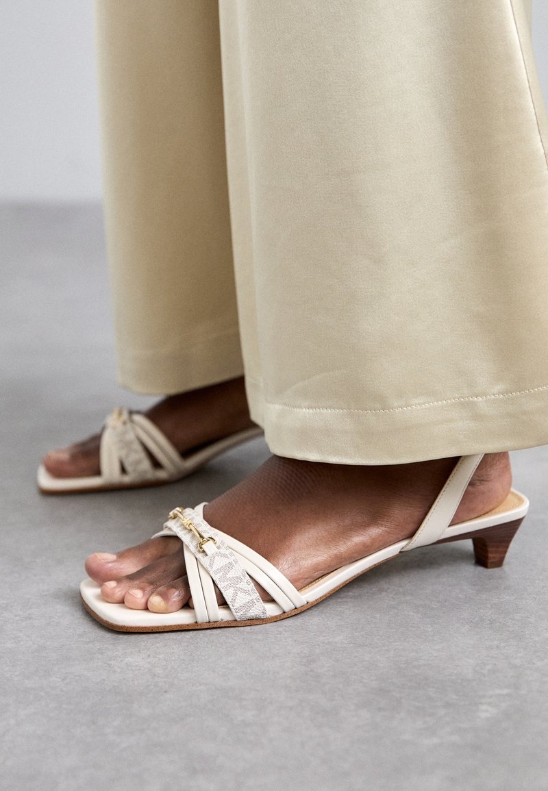Pieds en sandales à talons beiges avec des lanières décoratives, portant un pantalon large couleur crème clair, debout sur un sol gris.