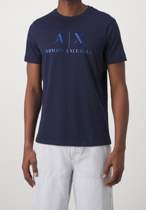 Mężczyzna w granatowej koszulce Armani Exchange z krótkim rękawem i niebieskim logo oraz białych spodniach, stojący na tle jednolitego jasnego tła.