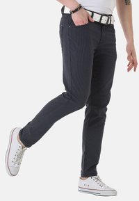 Dunkel gemusterte Jeans, schmal geschnitten, mit fünf Taschen, kombiniert mit einem weißen Gürtel und weißen Sneakers mit roten Akzenten.