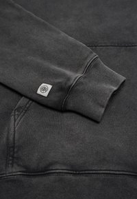 Tummanharmaa huppari, jossa on resorit hihansuissa ja etutasku, sekä pieni ommeltu merkki hihassa. Valmistettu pehmeästä, tekstuuripinnasta.