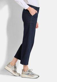 Pantalon en denim bleu foncé avec une coupe droite, comportant une poche avant et des coutures contrastées. Associé à des chaussures de sport gris clair et argent.