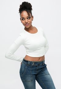 Fehér hosszú ujjú, rövid fazonú felső bordázott textúrával, sötétkék magasderekú farmerrel párosítva, elején zsebekkel és gombos záródással.