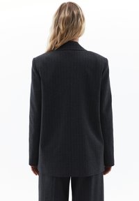 Mørk grå blazeren med stribede mønstre har et struktureret design, med udskårne revers og lange ærmer. Materialet synes at være tekstureret og skræddersyet.