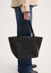 Borsa tote in pelle nera con superficie testurizzata, dotata di due manici robusti e un ampio appertura superiore. Indossata da una persona in jeans blu.