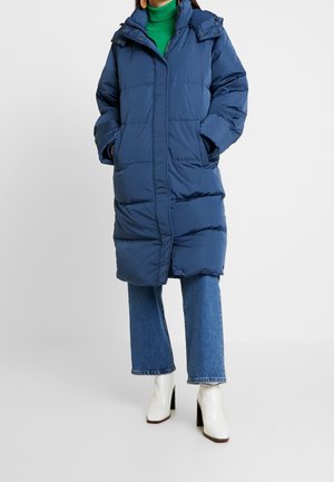 Femme portant un long manteau matelassé bleu marine sur un jean bleu et des bottines blanches à talons, debout les mains dans les poches du manteau.