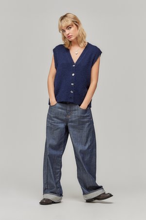 Blonde vrouw in een marineblauwe, knooploze mouwloze trui, wijde jeans met omslag en zwarte schoenen, met haar handen in de zakken tegen een effen achtergrond.