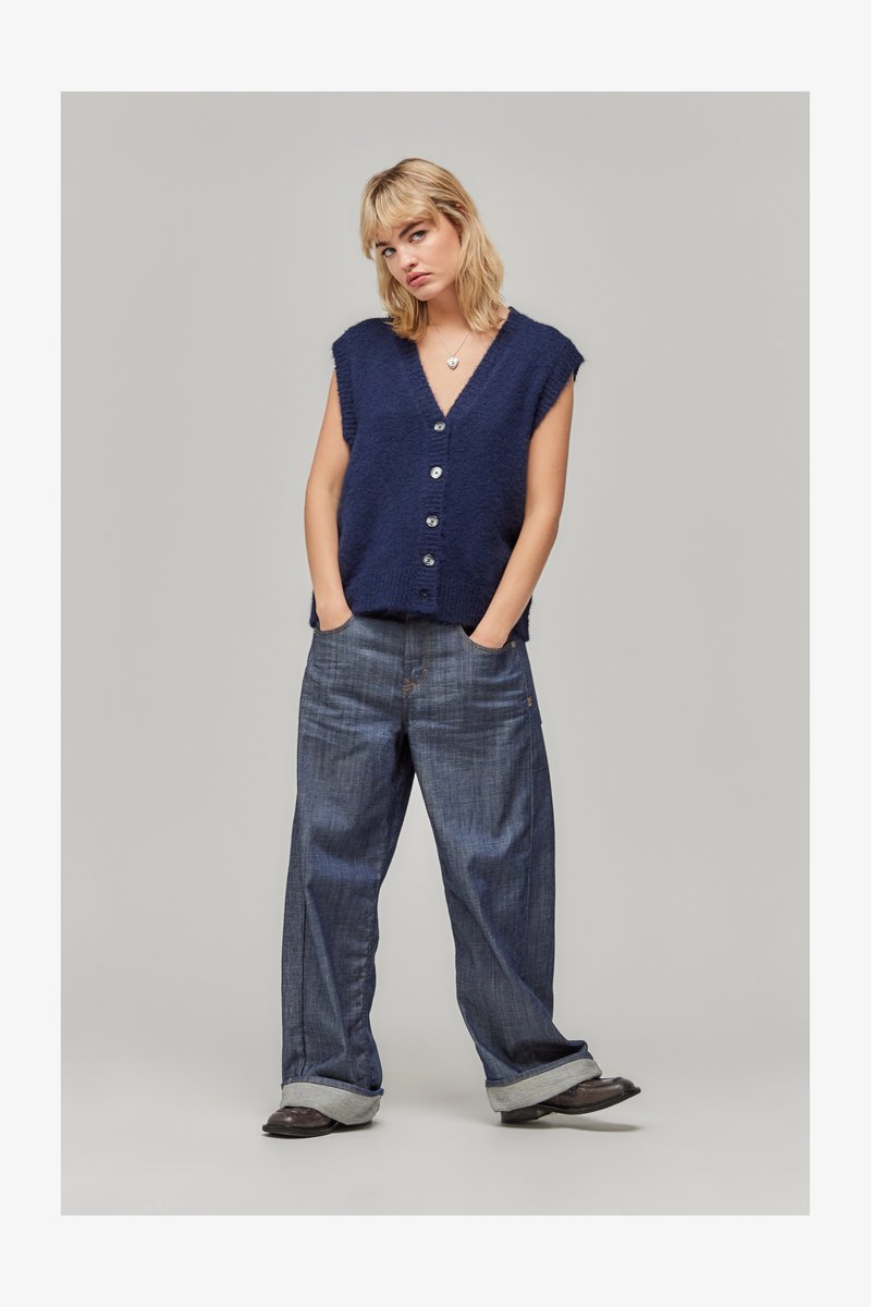 Ξανθιά γυναίκα φορώντας μια ναυτική μπλε αμάνικη μπλούζα με κουμπιά, φαρδιά τζιν με κουμπωμένα μανίκια, μαύρα παπούτσια, με τα χέρια στις τσέπες, μπροστά σε ένα απλό φόντο.