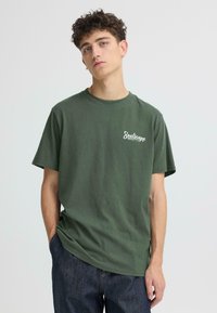 T-shirt en coton vert olive à manches courtes, col rond, avec logo blanc "Soulscape" sur la poitrine. Tissu texturé, coupe décontractée.
