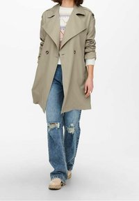 Trench-coat beige avec larges revers, coupe croisée à double boutonnage et détails à boutons ; associé à un jean bleu usé et des chaussures beiges.