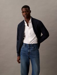 Marine bomberjack, witte overhemd, en blauwe denim jeans. Glad stof, klassieke snit, en contraststiksels op de jeans.