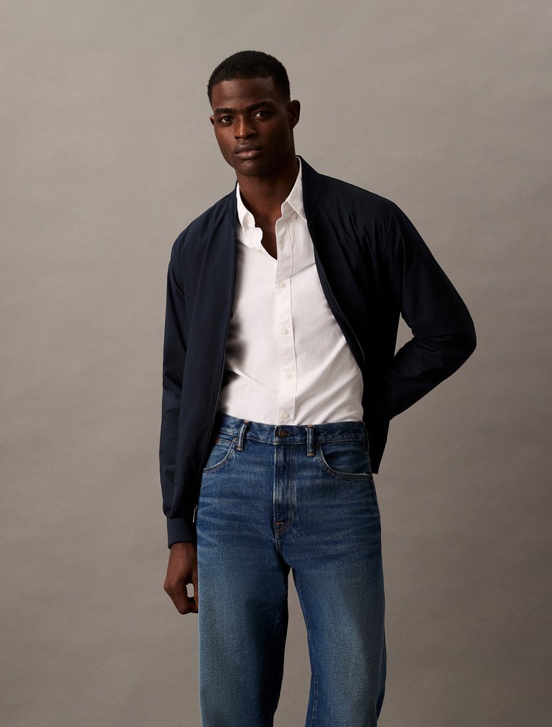 Marine bomberjack, witte overhemd, en blauwe denim jeans. Glad stof, klassieke snit, en contraststiksels op de jeans.