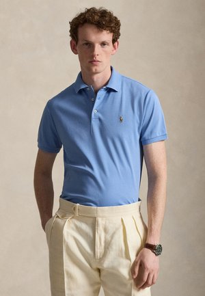 Młody mężczyzna z kręconymi włosami nosi jasnoniebieską koszulkę polo i kremowe plisowane spodnie, stojąc na tle gładkiego beżowego tła.