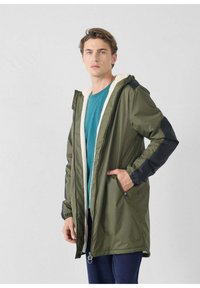 Olijfgroene, waterdichte parka met een crème kleurige, gevoerde capuchon van fleece, zwarte accenten op de mouwen, zakken met rits en een losse pasvorm.