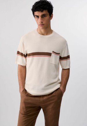 Giovane uomo con capelli ricci scuri che indossa una maglia beige a maniche corte lavorata a maglia con strisce orizzontali marroni e rosse e pantaloni marroni abbinati.