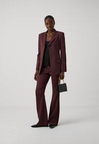 Traje entallado burdeos con pantalones acampanados y blazer de un solo botón, combinado con una blusa negra y un pequeño bolso negro.