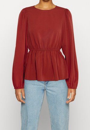 Femme portant un chemisier à manches longues couleur rouille avec taille froncée et manches bouffantes, associé à un jean bleu clair.