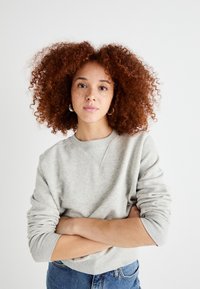Sweatshirt gris clair avec un col rond et des poignets côtelés, porté sur un jean bleu, associé à des boucles d'oreilles créoles et des cheveux bouclés brun rougeâtre.