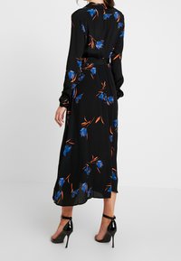 Robe noire à manches longues, motif floral en bleu et orange, taille ceinturée, tissu fluide, longueur au niveau des chevilles avec des chaussures noires à talons.
