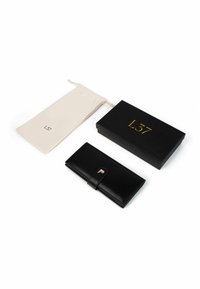 Czarny skórzany portfel z ozdobą ze złotego metalu, w zestawie z kremowym woreczkiem na sznurek i czarnym pudełkiem z złotym napisem.