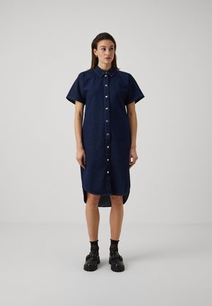Navy denim shirt dress with short sleeves, front button closure, and a curved hem. Paired with black platform shoes and dotted socks.