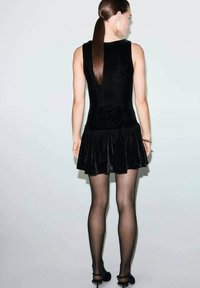 Kobieta z długimi prostymi włosami w czarnej bezrękawnikowej aksamitnej sukience z rozkloszowanym dołem, przezroczystych czarnych rajstopach i czarnych sandałach na obcasie, stojąca bokiem.