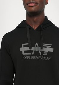 Črna kapuca z vrvico, z velikim sivo grafičnim logotipom, ki na sprednji strani prikazuje "EA" in "EMPORIO ARMANI". Mehka tekstura.