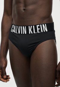 Ανδρικός κορμός που φοράει μαύρα εσώρουχα Calvin Klein με φαρδιά λωρίδα με λογότυπο, σε λευκό απλό φόντο.
