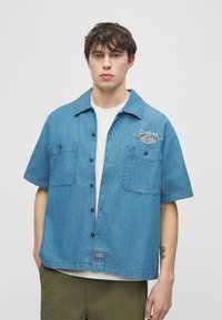 Νεαρός άνδρας που φοράει μπλε κοντομάνικο τζιν πουκάμισο με μπροστινές τσέπες και το λογότυπο "Dickies" πάνω από λευκή κοντομάνικη μπλούζα και χακί παντελόνι.