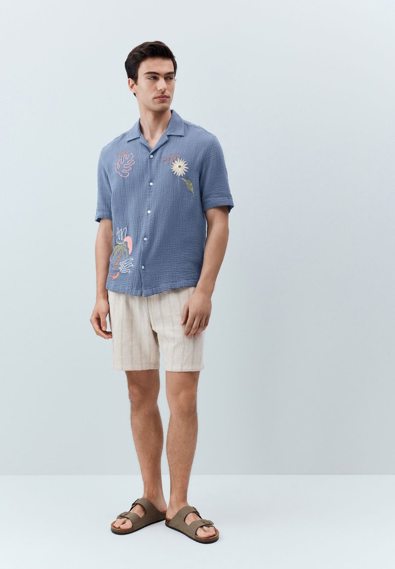 Giovane uomo in piedi, indossa una camicia blu a maniche corte con ricamo floreale, pantaloncini beige e sandali marroni su uno sfondo semplice.