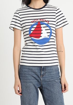 Kobieta nosząca białą i granatową koszulkę w paski z czerwono-białą grafiką żaglówki i niebieskim kołem, w połączeniu z niebieskimi dżinsami.