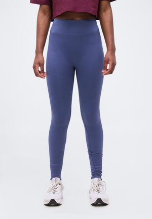 Γυναίκα σε μπλε κολάν με ψηλή μέση και λευκά αθλητικά παπούτσια, σε όρθια στάση με τα χέρια χαλαρά στα πλάγια, μπροστά σε απλό φόντο.