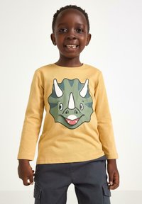Smilende dreng iført en gul langærmet skjorte med ansigtet af en grøn triceratops, der har en lynlåslukket mund, og mørke cargo-bukser.