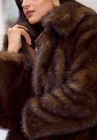 Rudas dirbtinio kailio paltas su minkštu, tekstūriniu paviršiumi, turintis užtrauktuką ir plačias rankoves. Artimas vaizdas, akcentuojantis purų audinį.